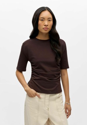 Femme aux longs cheveux foncés portant un t-shirt marron foncé à manches courtes et un pantalon beige, debout avec une main dans la poche sur un fond blanc.