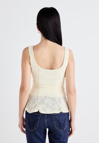 Model in een lichtgele peplum top met kantdetails, gecombineerd met blauwe jeans, gezien vanaf de achterkant tegen een effen achtergrond.