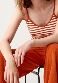 Pruhovaný pletený top na ramínka v oranžové a krémové barvě, s výstřihem do V a tenkými ramínky, spárovaný s oranžovými širokými kalhotami, které mají strukturovanou tkaninu.