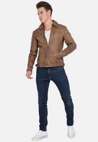 Giubbotto di pelle marrone con chiusura a zip, colletto a punta e design aderente, abbinato a jeans blu scuro e sneaker bianche.