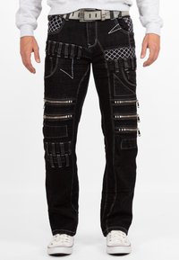 Jeans in denim nero con più cerniere, modelli di cucitura unici e accenti texturizzati. Include una cintura grigia e sneakers bianche casual.