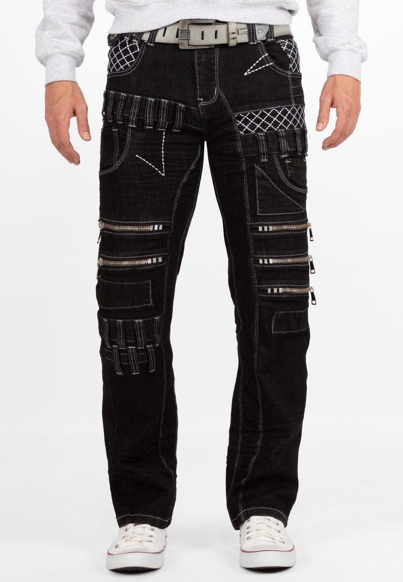 Jeans in denim nero con più cerniere, modelli di cucitura unici e accenti texturizzati. Include una cintura grigia e sneakers bianche casual.