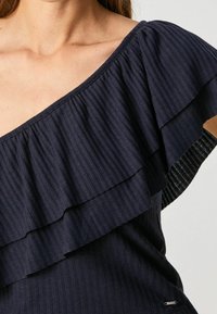 Tmavě modrý top s ribbovanou tkaninou a s vrstvami volánů na levém rameni. Obsahuje malé logo na dolním pravém rohu.