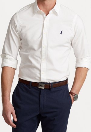 Man draagt een wit overhemd met knopen en opgerolde mouwen, marineblauwe broek, bruine leren riem en een polshorloge met een bruine band.