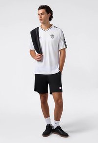 Wit atletisch shirt met zwarte accenten, v-hals ontwerp en logo; gecombineerd met zwarte shorts en zwarte sneakers. Zwarte handdoek over de schouder gehangen.