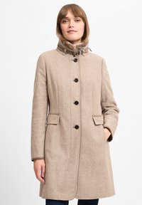 Beige uldfrakke med høj krave, knaplukning og sidelommer. Har et struktureret herringbone-mønster og kontrasterende syninger.