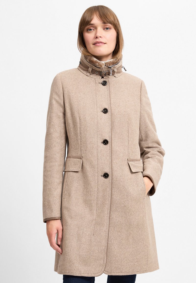 Beige uldfrakke med høj krave, knaplukning og sidelommer. Har et struktureret herringbone-mønster og kontrasterende syninger.