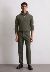 Olivgrüner Strickpullover mit einem Reißverschlusskragen, kombiniert mit passenden maßgeschneiderten Hosen und schwarzen Lederschuhen, vor einem neutralen Hintergrund stehend.