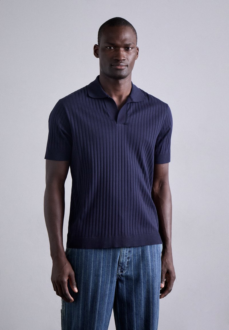 Polo en tricot côtelé bleu marine avec un petit col et des manches courtes, associé à un jean rayé bleu clair.