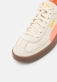 Baskets Puma avec un dessus crème, en suède et cuir, semelle en caoutchouc texturé couleur tan et accents roses sur le côté.