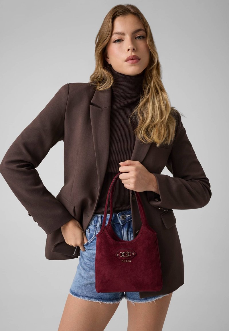 Femme portant un blazer marron et un col roulé noir, tenant un sac à main rouge Guess et portant un short en jean sur un fond uni.