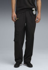 Schwarze Cargo-Hosen mit elastischem Bund, seitlichen Taschen und strukturiertem Stoff. Grünes Logodetail auf der oberen linken Seite.