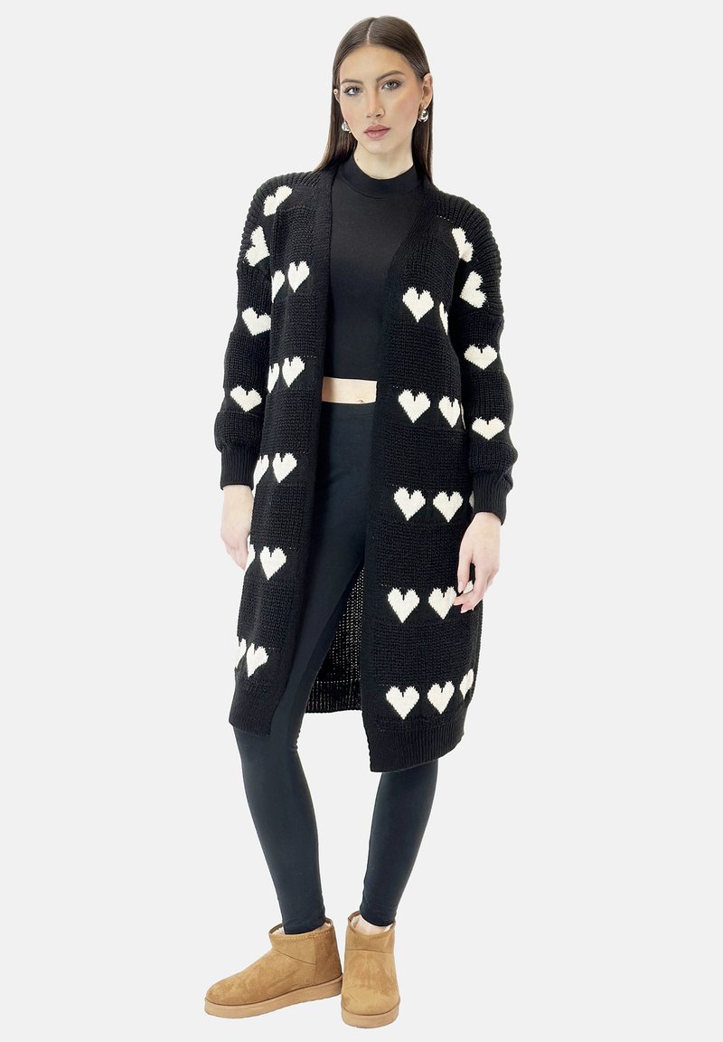 Zwarte gebreide cardigan met witte hartpatronen, open voorkant, lange mouwen en geribbelde manchetten, gecombineerd met een zwarte top en leggings.