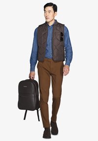 Gilet imbottito marrone, camicia in denim blu, pantaloni marroni e scarpe scure. Porta uno zaino marrone scuro con una texture liscia e dettagli con cerniera.