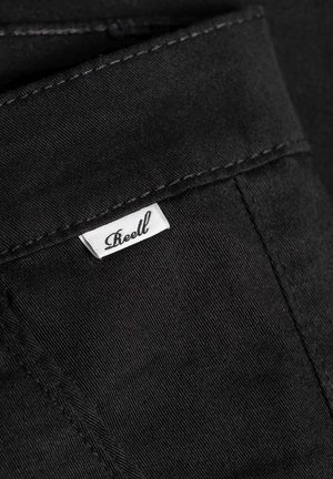Tela de mezclilla negra con una textura sutil, con una etiqueta blanca cosida en el bolsillo que dice "Reell". Las costuras oscuras contrastan con el material.