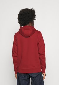 Persona con cabello rizado que lleva una sudadera roja simple y vaqueros azul oscuro, vista desde atrás contra un fondo claro y liso.