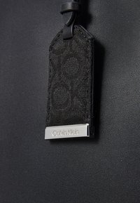 Black synthetic bag with a textured woven tag featuring a geometric pattern and a metallic silver label inscribed with "Calvin Klein."
