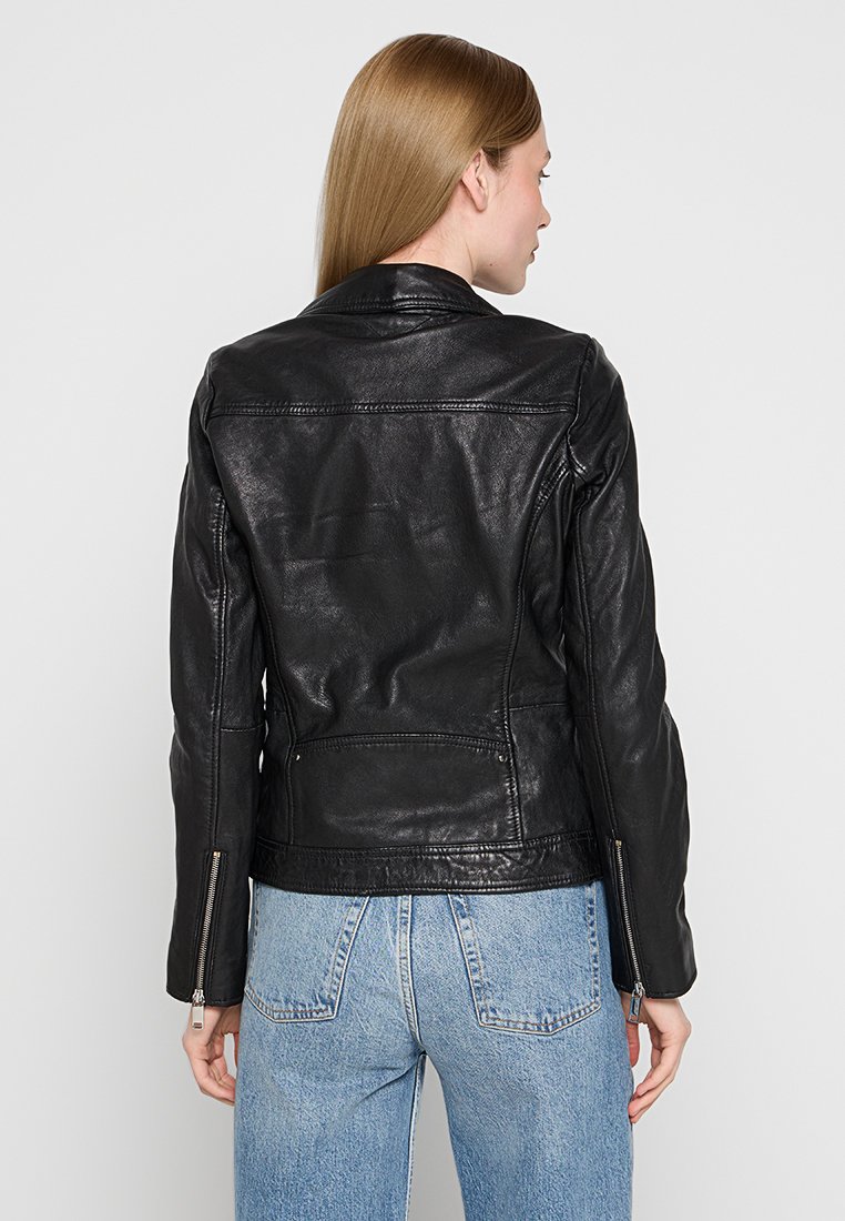 Giubbotto in pelle nera da motociclista con design aderente, cuciture a vista e polsini con zip. Indossato sopra jeans in denim chiaro.