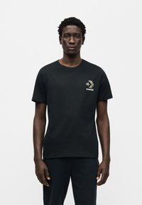 Μαύρο βαμβακερό t-shirt με κοντά μανίκια, που διαθέτει ένα μικρό πράσινο και κίτρινο σχέδιο με φύλλα και τη λέξη "Converse" στο αριστερό στήθος.