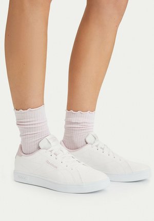 Ben iført lyserøde ribstrikkede sokker og hvide Reebok-sneakers med snørebånd, fotograferet mod en ensfarvet hvid baggrund.