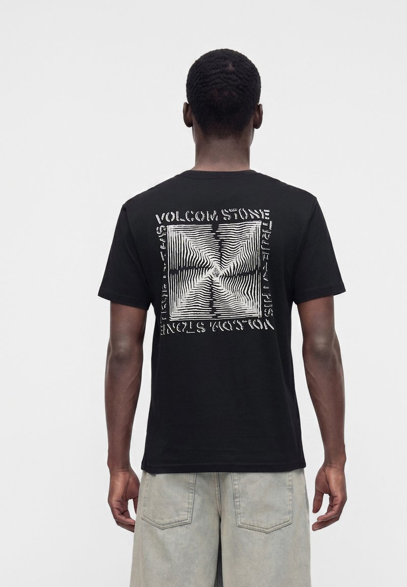 Homme portant un t-shirt noir à manches courtes avec un motif carré abstrait blanc et un texte dans le dos, associé à un short de couleur claire.