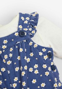 Blaue Baumwoll-Latzhose mit weißen Gänseblümchenmustern, rüschigen Trägern, dunklen Knöpfen und einem cremefarbenen Langarmshirt darunter.