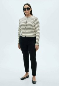 Gestreifter cremefarbener Cardigan mit schwarzen Linien, schwarze eng anliegende Hose und schwarze Ballerinas mit Akzenten. Das Model trägt große Sonnenbrillen und hat die Haare offen.