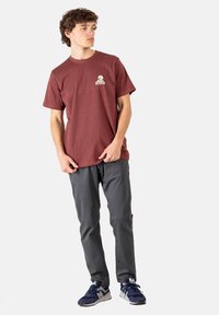 Camiseta de algodón color burdeos con mangas cortas y un logo. Combinada con pantalones grises y zapatillas oscuras. Modelo de pie con las manos en los bolsillos.