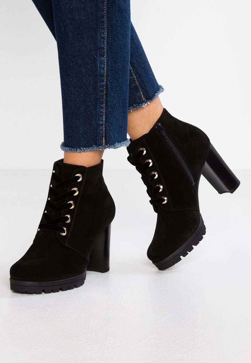 Bottes noires en daim à talons hauts et à la cheville, avec œillets dorés et fermeture éclair latérale, portées avec un jean bleu foncé à ourlet effiloché sur un sol blanc.