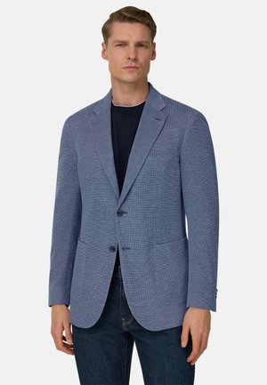 Homme portant un blazer bleu texturé sur une chemise noire et un jean foncé, debout devant un fond clair uni.