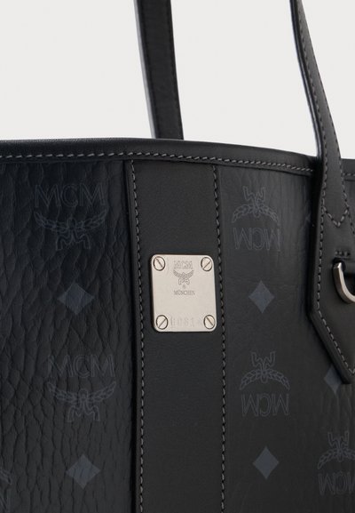 Gros plan sur un sac cabas MCM en cuir noir avec des logos embossés et une plaque métallique argentée indiquant "MCM München" et un numéro de série.