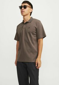 Hnedé polo tričko s krátkym rukávom a golierom, s malým vyšívaným logom na ľavej hrudi. Nosené s tmavými nohavicami a slnečnými okuliarmi.