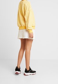 Persona con un suéter a rayas amarillas y blancas, una mini falda blanca deshilachada y zapatillas gruesas en blanco y negro, de pie sobre un fondo liso.