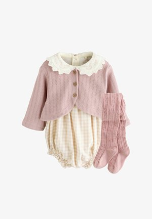 Pull en tricot rose clair avec un col en dentelle blanche et des boutons en bois, associé à une combinaison à carreaux beige et des chaussettes hautes roses assorties.