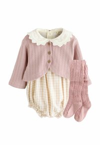 Jasnoróżowy sweter z dzianiny z białym koronkowym kołnierzykiem i drewnianymi guzikami, w połączeniu z beżowym kombinezonem w kratę i pasującymi różowymi skarpetkami do kolan.