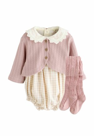 Helle rosa gestrickte Sweater mit einem weißen Spitzenkragen und Holzknöpfen, kombiniert mit einem beigen karierten Overall und passenden rosa kniehohen Socken.