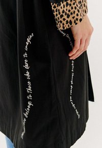 Čierna látková bunda s rukávom so leopardím vzorom. Obsahuje biely text pozdĺž lemu, má hladkú textúru a voľný strih.