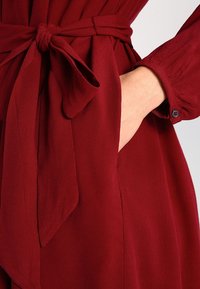 Burgundia mekko, jossa on solmittava vyötärö, pitkät hihat ja sivutasku. Valmistettu pehmeästä, virtaavasta kankaasta, jolla on sileä rakenne.