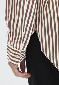 Stribet skjorte i creme og brun, med knapmanchet og forlænget bagkant. Stoffet ser glat ud med en let glans.