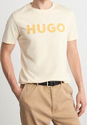 Mand iført cremefarvet T-shirt med korte ærmer og stort gult "HUGO"-tryk, stoppet ned i beige bukser med sort bælte.
