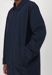 Mørkeblå lang frakke, lavet af letvægtsmateriale, med spids krave, sidelommer og en subtil logoaccent på ærmet.