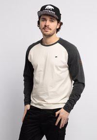 Long-sleeve T-shirt featuring a cream body and dark gray raglan sleeves, with a small bear logo on the chest and brown accent on the sleeve.