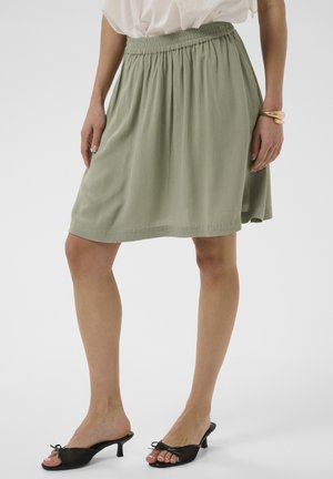 Frau trägt einen hellgrünen knielangen Rock mit elastischem Bund, kombiniert mit schwarzen, niedrighackigen offenen Schuhen und einem goldenen Armband.