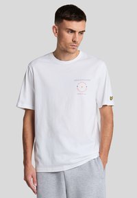 Camiseta blanca de algodón con cuello redondo, mangas cortas y un gráfico impreso en naranja y gris en el lado izquierdo del pecho. Acento de logo amarillo en la manga.