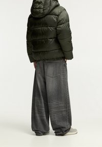 Chaqueta de plumas verde con capucha, combinada con unos vaqueros de mezclilla gris de corte oversized. La chaqueta presenta secciones acolchadas; los vaqueros tienen un patrón desgastado.