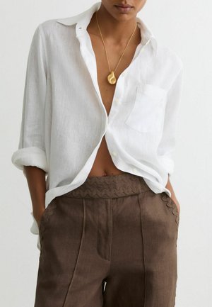 Person trägt ein weißes Hemd mit Knöpfen und hochgekrempelten Ärmeln, braune Hose mit wellenförmigem Besatz und eine goldene Anhängerhalskette. Hände in den Taschen.