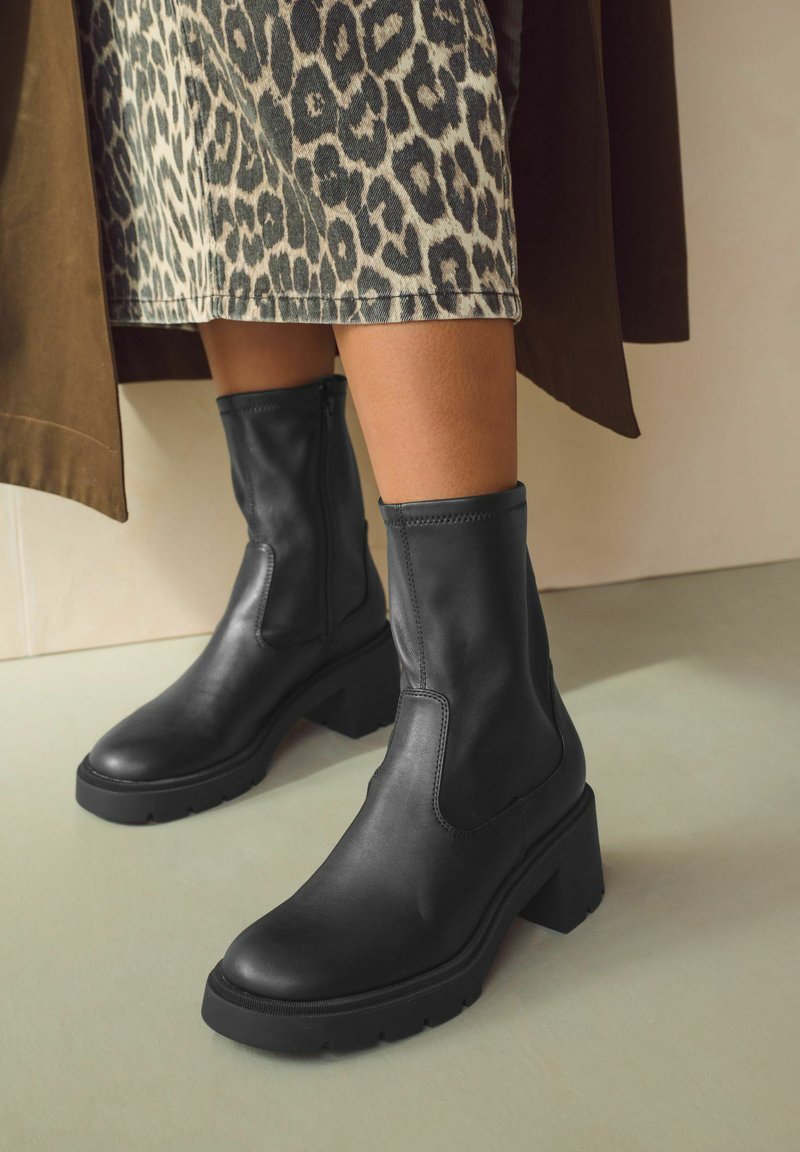 Black leather ankle boots with elastic side panels and a chunky block heel. The soles are textured for grip, suitable for various surfaces.