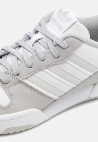 Zapatillas deportivas de cuero gris con acentos blancos, que cuentan con tres rayas blancas, cordones planos y una lengua acolchada con un detalle de logo.
