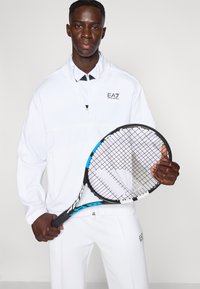 Λευκό αθλητικό μπουφάν και παντελόνι με το λογότυπο EA7, συνδυασμένα με μαύρο και μπλε ρακέτα τένις, που διαθέτει μοτίβο πλέγματος στα χορδές.