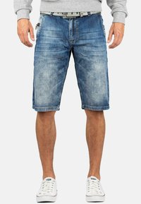 Shorts en denim bleu avec un délavage, fermeture éclair et bouton, poches avant et une ceinture grise. Associés à des baskets blanches.